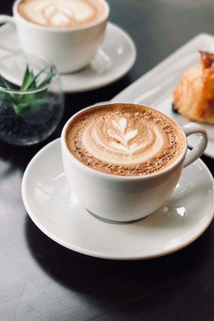 A close-up of a cup of coffee with latte art on a wooden café table, with another coffee cup and pastry in the background.